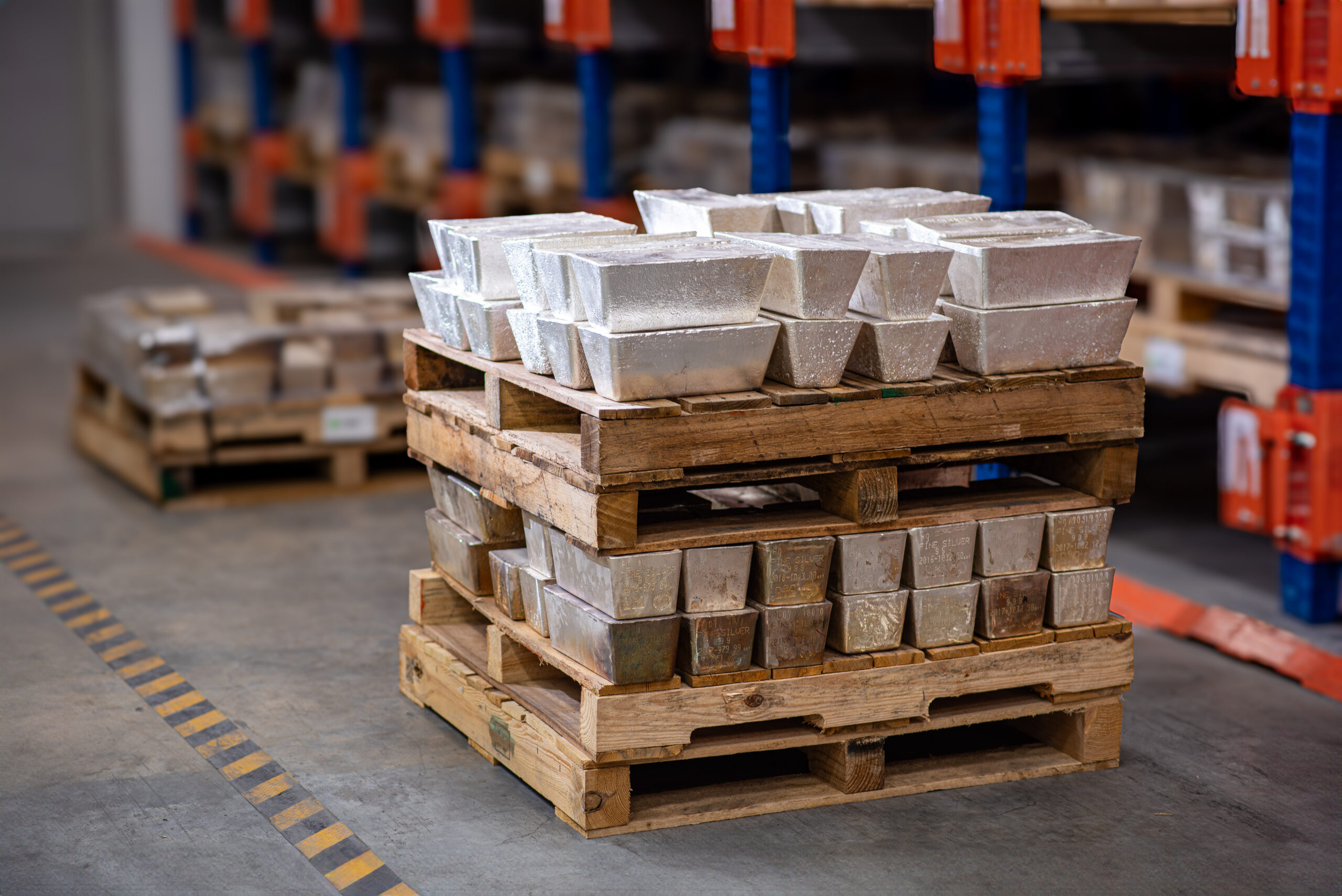 Artistic shallow depth of field shot of 1000 oz silver bars stacked on stacked pallets, sitting on the floor of a depository facility. Background includes racking system and an out of focus pallet.